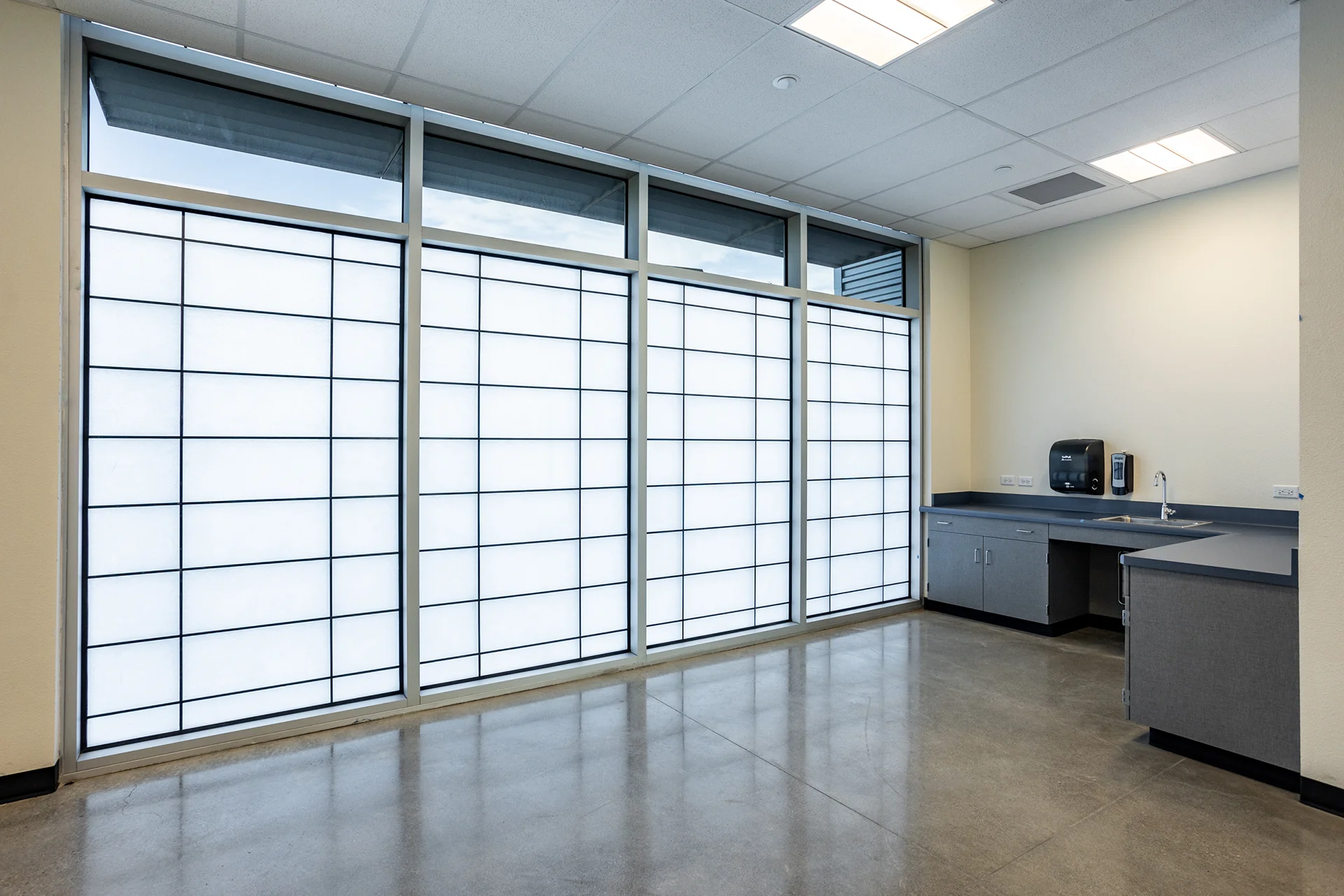 School faculty work room renovation showing a multi-panel gridded translucent window wall, gray base cabinetry with a stainless steel utility sink, automatic soap dispensers, polished concrete floors, and a solid plaster wall backdrop.
