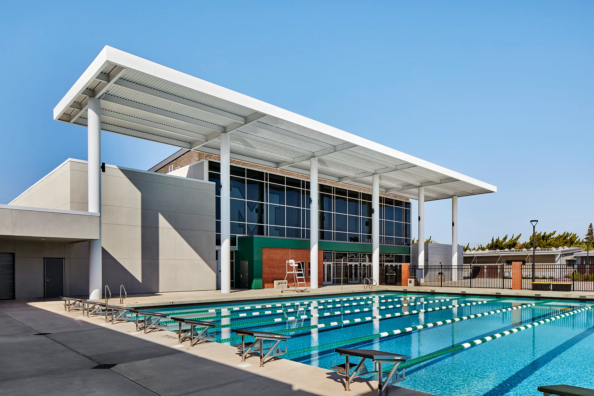 Clean daytime view of a public school competitive swimming pool installation, showcasing clear blue water, a lifeguard tower station, a white-coated structural steel open-air pavilion ceiling, and a brick masonry facade transition leading into the indoor fitness wings.