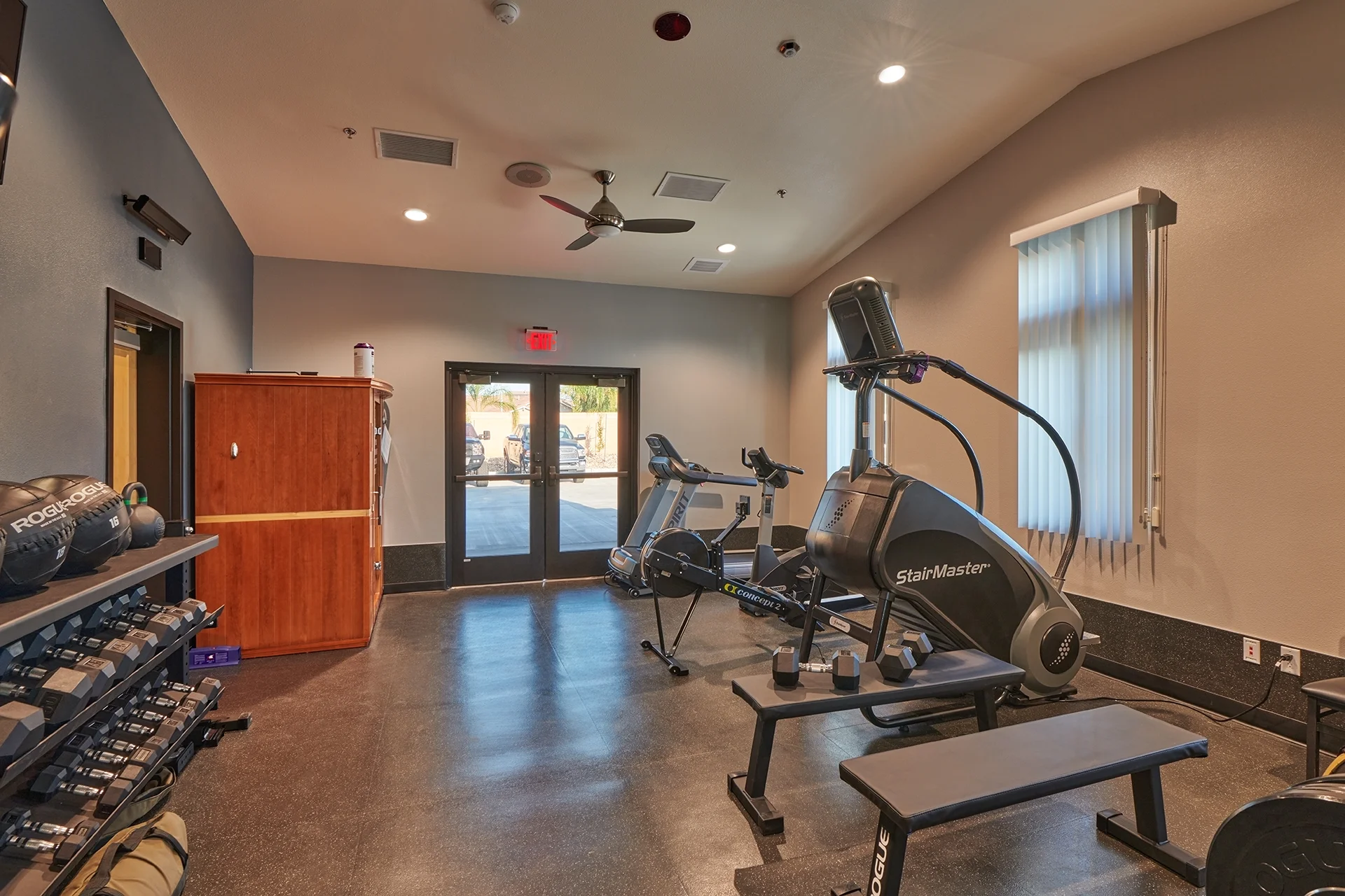 Specialized station exercise room featuring a StairMaster machine, a stationary training bike, free weight benches, a dumbbell storage rack, an infrared carcinogen mitigation sauna booth, and large glass exit doors.