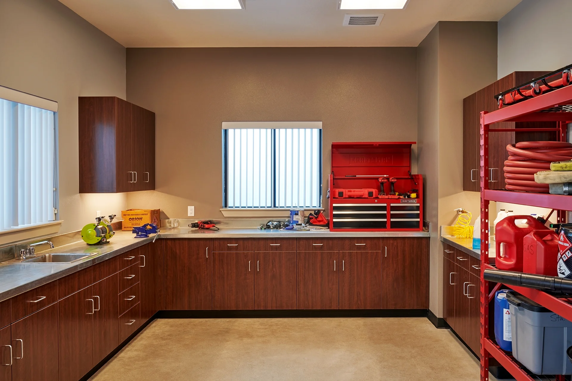 Interior of the station maintenance workshop room featuring a sealed concrete floor, dark wood tool storage cabinetry, continuous stainless steel countertops with an integrated utility sink, and a red metal Craftsman tool chest.