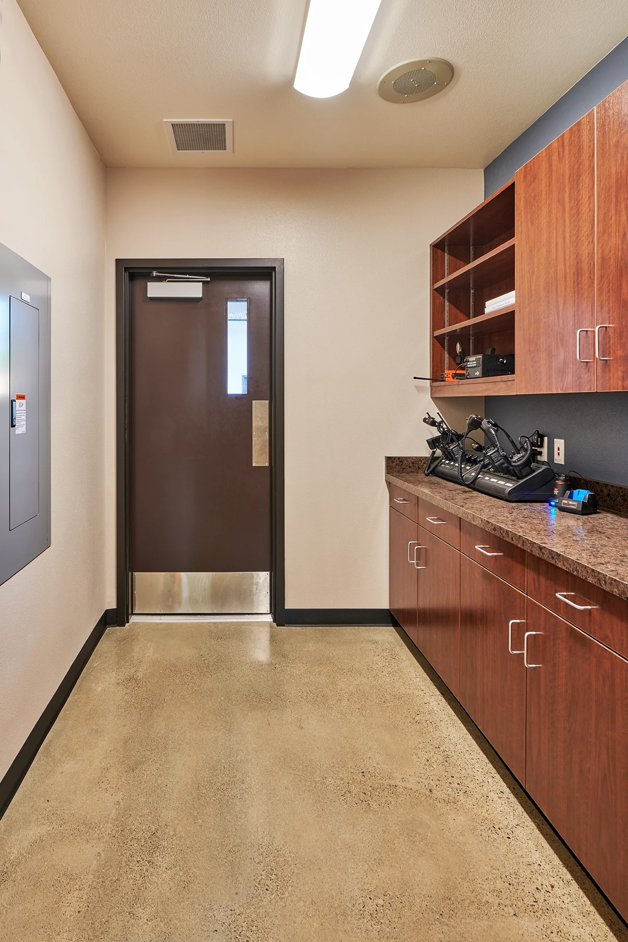 Interior service corridor with stained concrete flooring, featuring a heavy-duty dark brown exit door with a kickplate, a wall-mounted electrical panel, and custom wood cabinetry supporting multi-unit radio and equipment charging docks.