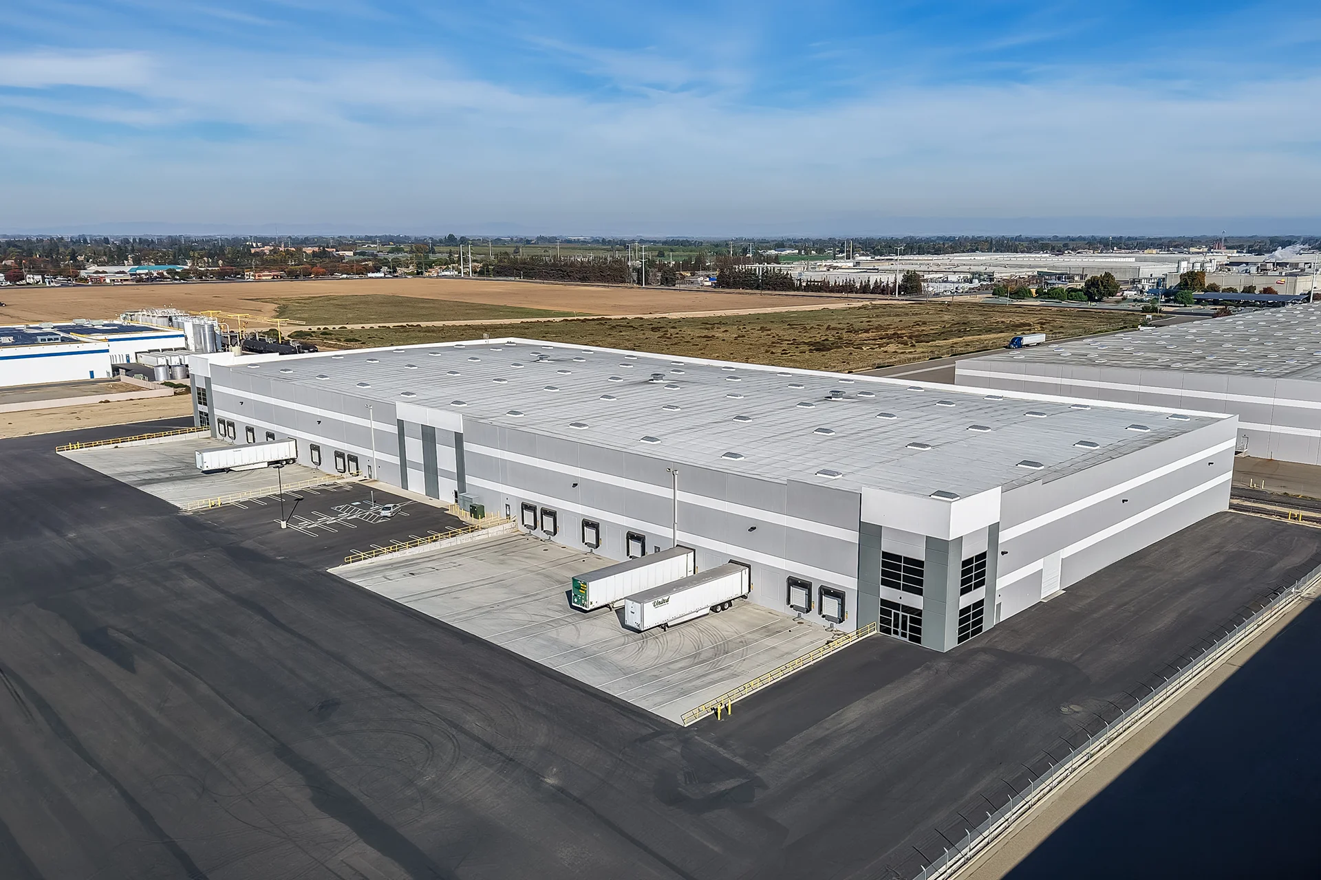 Angular aerial overview of a completed concrete tilt-up warehouse asset, emphasizing structured roof drainage lines, multiple active logistics trailer units at cross-dock positions, concrete dolly pads, and a continuous chain-link security perimeter fence.