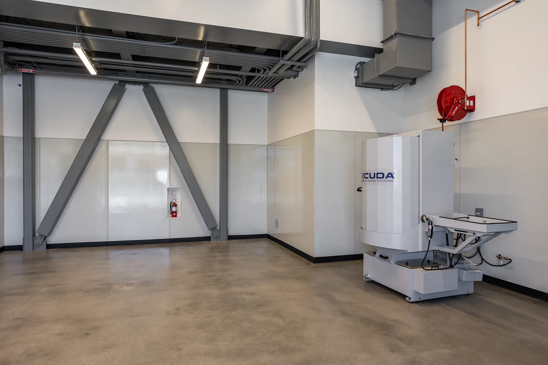 Open-concept industrial workshop interior highlighting exposed V-shaped structural steel seismic bracing beams, smooth concrete flooring, overhead conduit tracking, and a recess-mounted fire extinguisher cabinet next to specialized cleaning machinery.