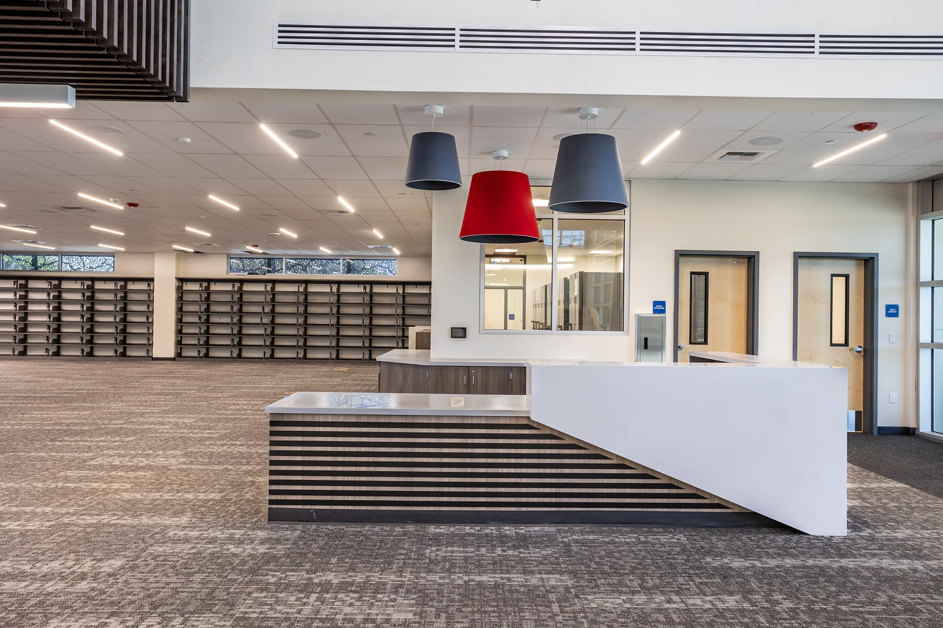 Interior shot of an institutional student services lobby featuring a modern angular customer service desk, linear ceiling strip lights, multi-toned decorative hanging shades, and clear view interior window partitions.