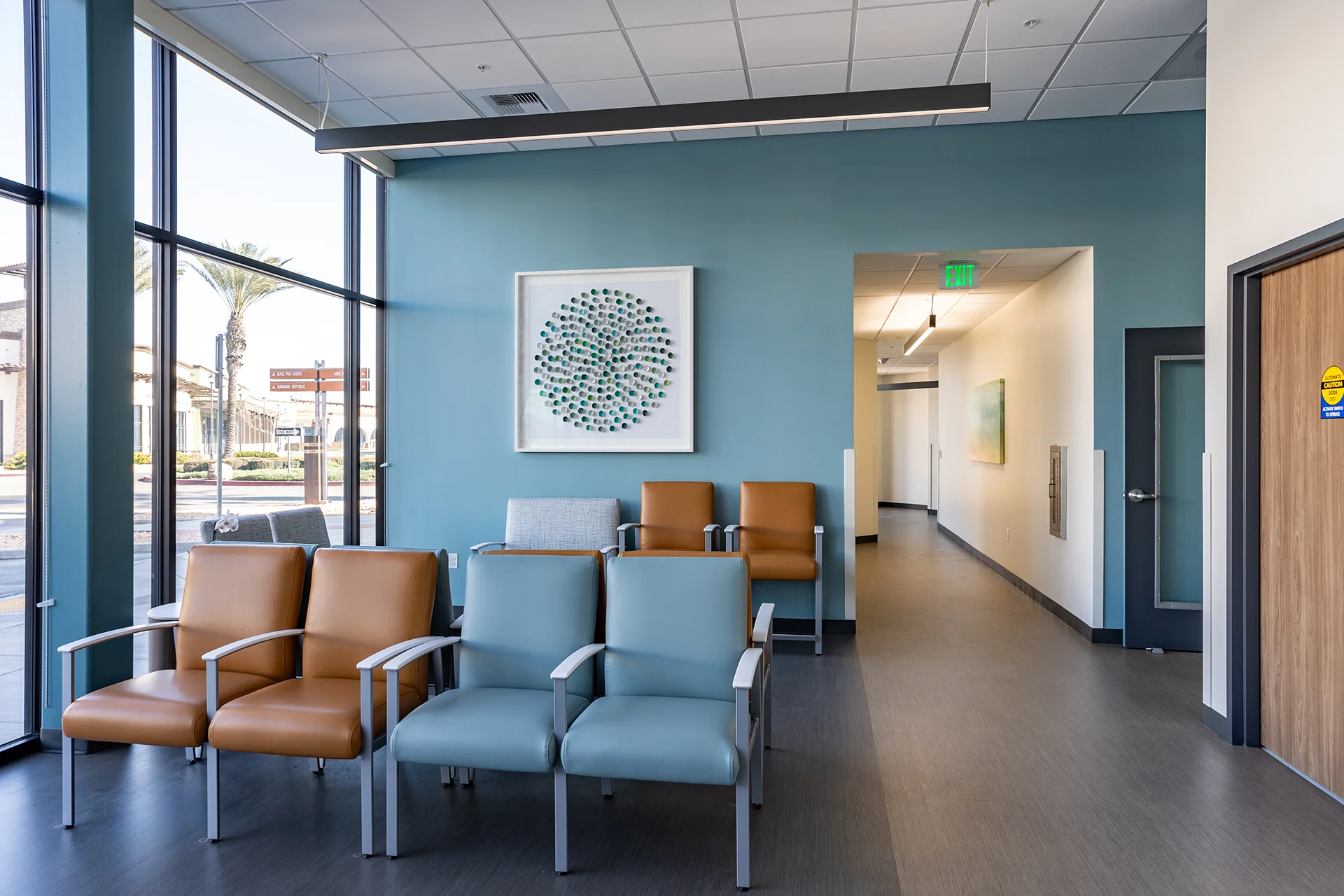 Medical office waiting area with panoramic storefront glass windows, featuring modular healthcare-grade armchairs in tan and sky blue vinyl, a wall-mounted circular mosaic art piece, a teal accent wall, and a linear pendant light fixture.
