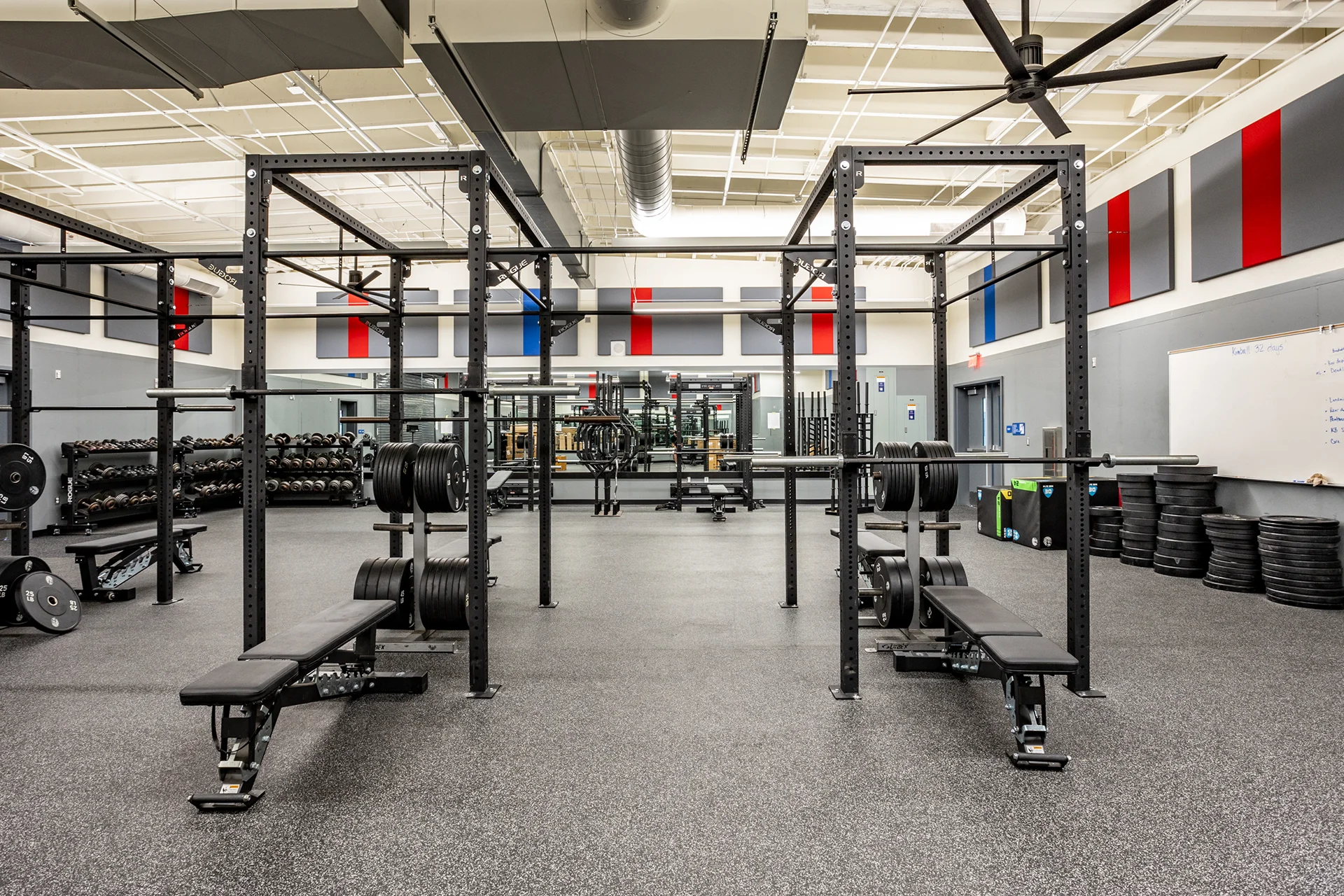 Symmetrical view of an institutional high school strength facility renovation showcasing parallel steel lifting cages, bumper plate stacks, industrial open ceiling ventilation ducts, and high-impact recycled rubber tile floors.