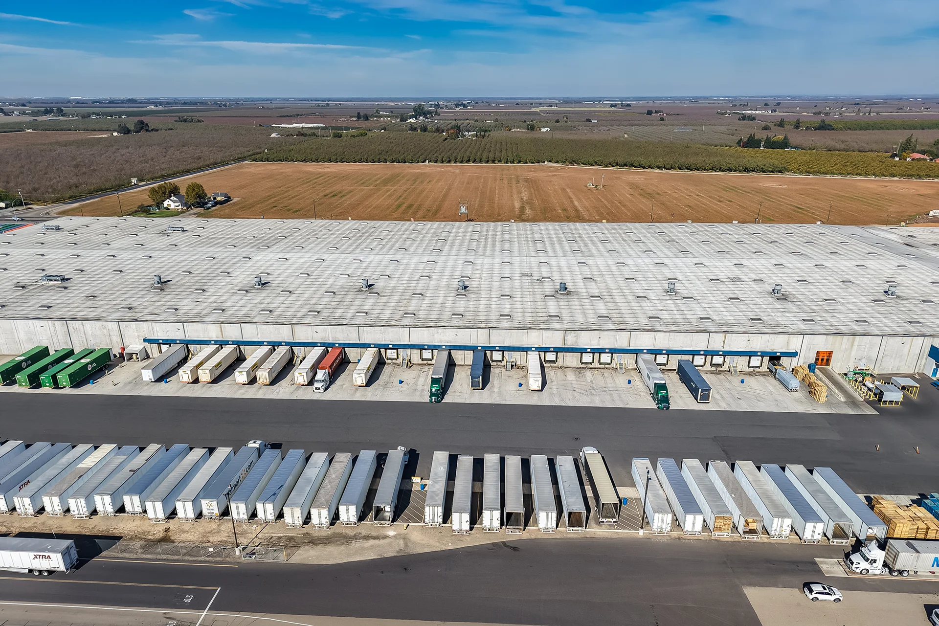 Longitudinal aerial view capturing the extensive cross-dock loading platform of a modern supply chain facility, featuring a structured line of insulated sectional overhead doors, heavy-duty rubber dock bumpers, organized rows of dropped trailers, and an expansive asphalt freight yard.