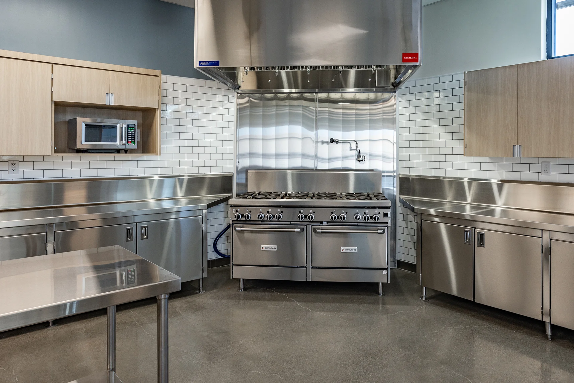 Balanced view of a commercial training kitchen showing dual stainless steel counter runs, an open utensil storage island, built-in culinary appliances, and structured white ceramic tile wall finishes under drop-ceiling troffer lights.