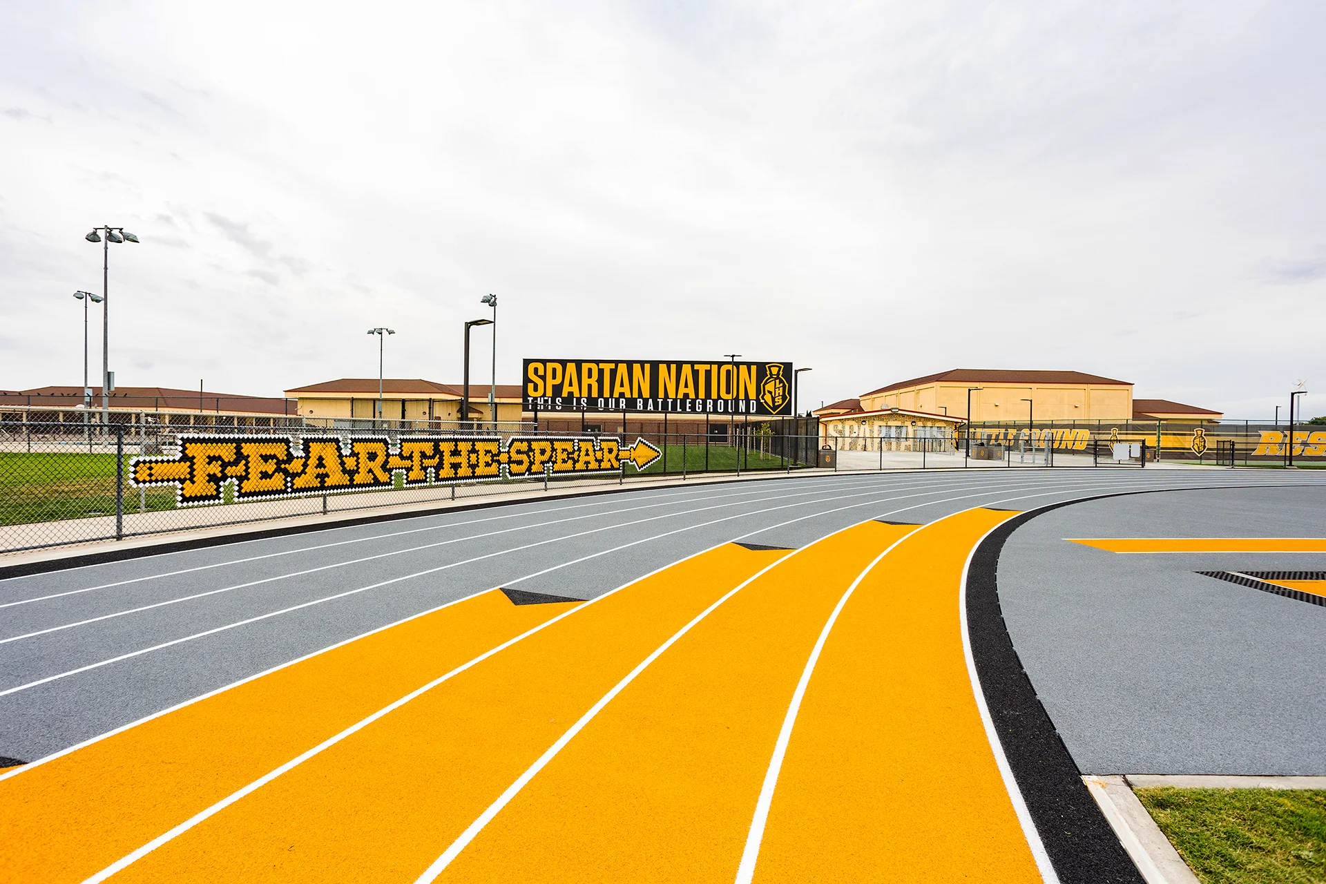 Ground-level view of a blue and yellow rubberized running track curve with a custom Spartan Nation sign on the perimeter fence.