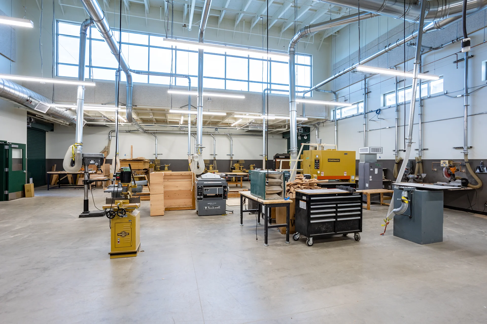 Full view of a modern school vocational technical shop room in San Joaquin County, highlighting stationary woodworking equipment, mobile tool chests, sheet material storage, and extensive galvanized steel dust collection pipes routed across white walls beneath a high ceiling with daylighting windows.