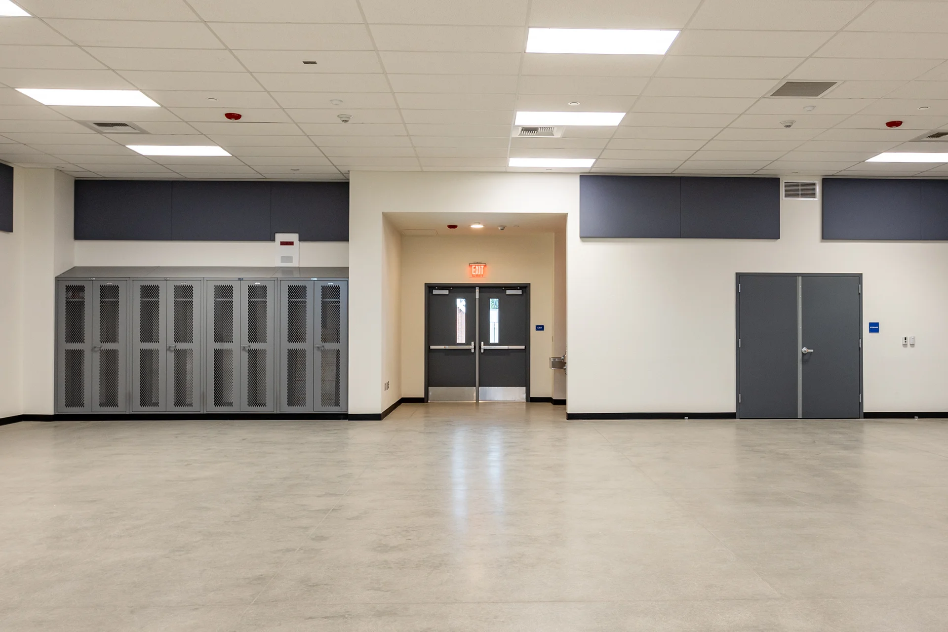Sports facility multi-purpose room showing a polished concrete floor, gray mesh-door storage lockers, a recessed stainless steel drinking fountain basin, and a dual door exterior emergency exit under an illuminated red sign.