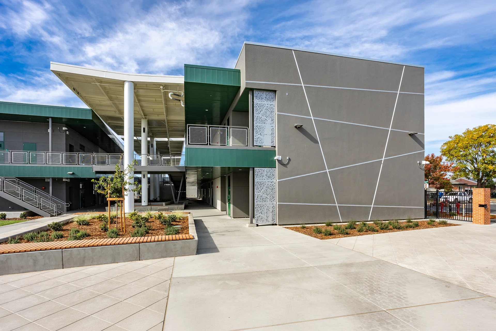 Ground-level perspective of a contemporary educational facility in Manteca, California, showcasing gray stucco facades with abstract white accent lines, decorative perforated metal privacy panels, an integrated concrete planter with wood slat seating, and elevated steel-frame walkways.