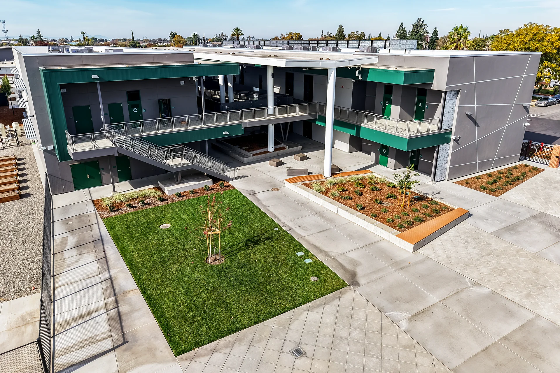 Symmetrical high-angle shot focusing on the central courtyard architecture of a contemporary school facility in Manteca, California, highlighting large white cylindrical support pillars, shaded lower plazas, tiered accent reveals on plaster walls, and a central lawn panel.