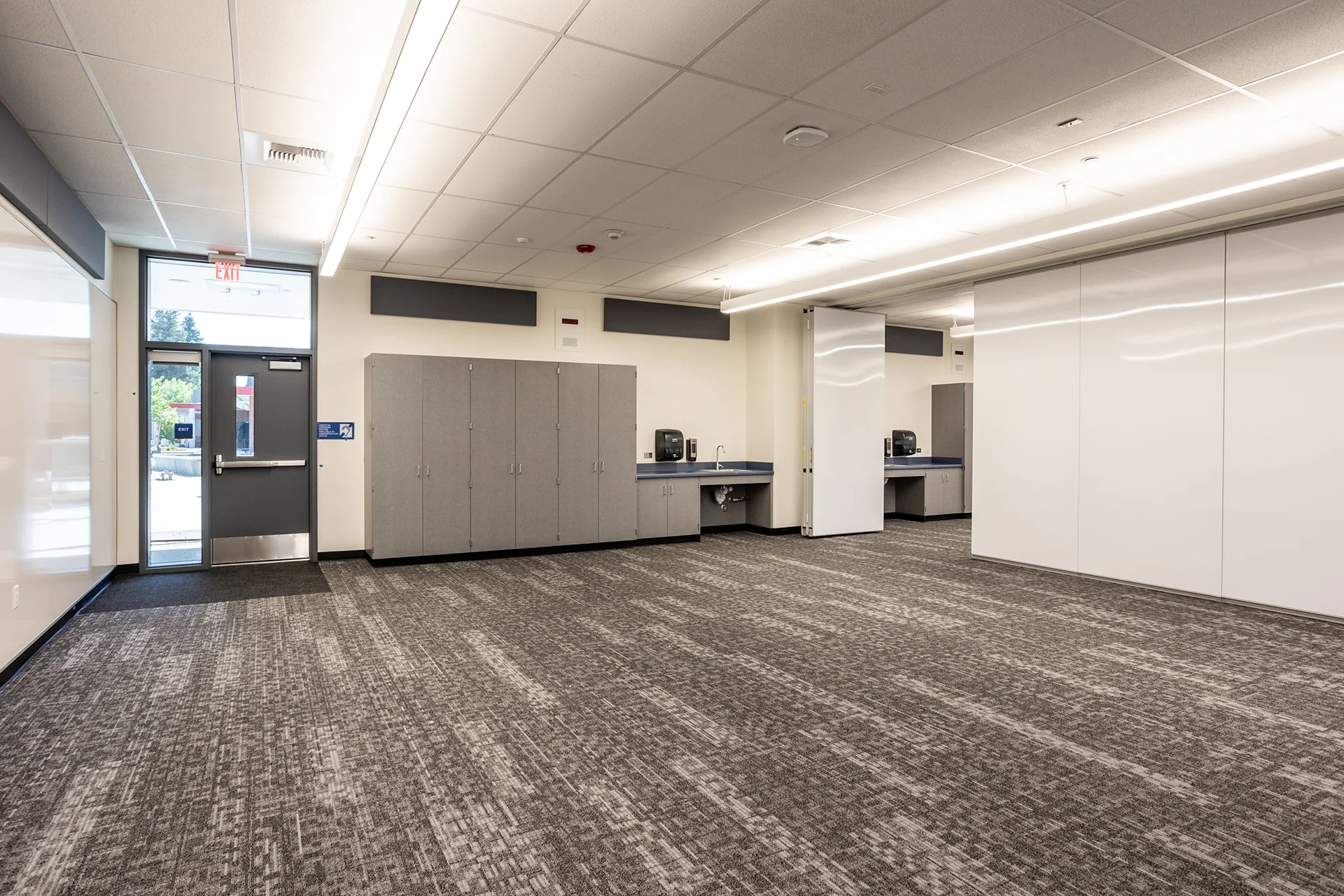 Academic wing common area featuring commercial-grade gray carpeting, matching laminate supply storage cabinets, integrated hand-washing sinks, and a large white sliding divider partition panel.