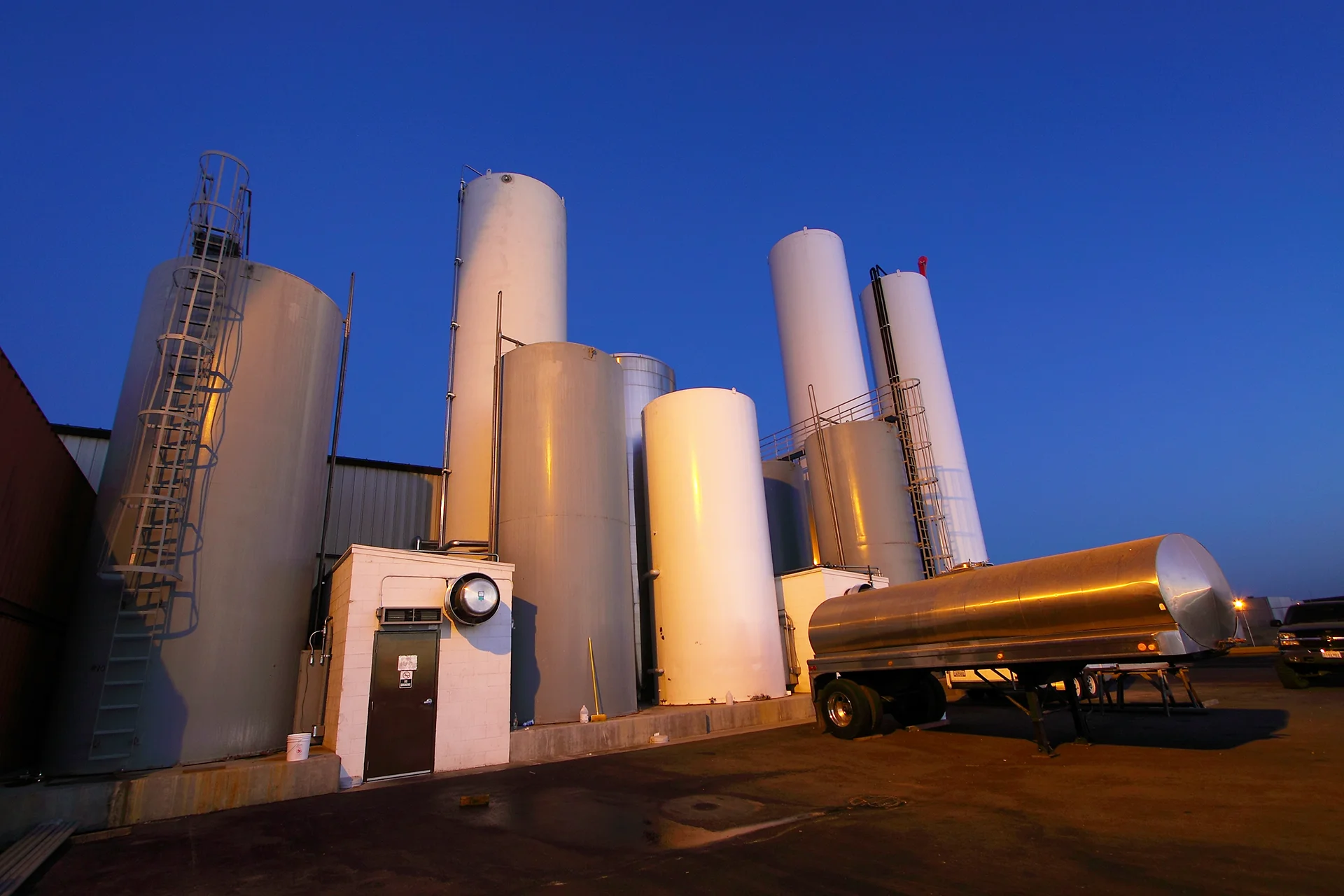 Angled view of a food-grade production building exterior detailing vertical bulk tanks of various diameters with safety cage ladders, a structural concrete containment foundation wall, a small concrete utility blockhouse, and a stainless steel bulk transport trailer.