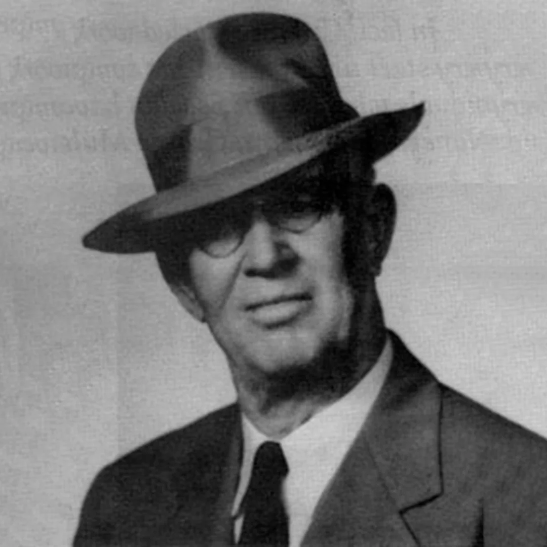 Vintage Portrait of Construction Company Founder Historic black-and-white studio portrait of an older gentleman wearing a classic fedora hat, eyeglasses, a dark suit jacket, and a tie, representing the heritage of a multigenerational family-owned general contracting firm.
