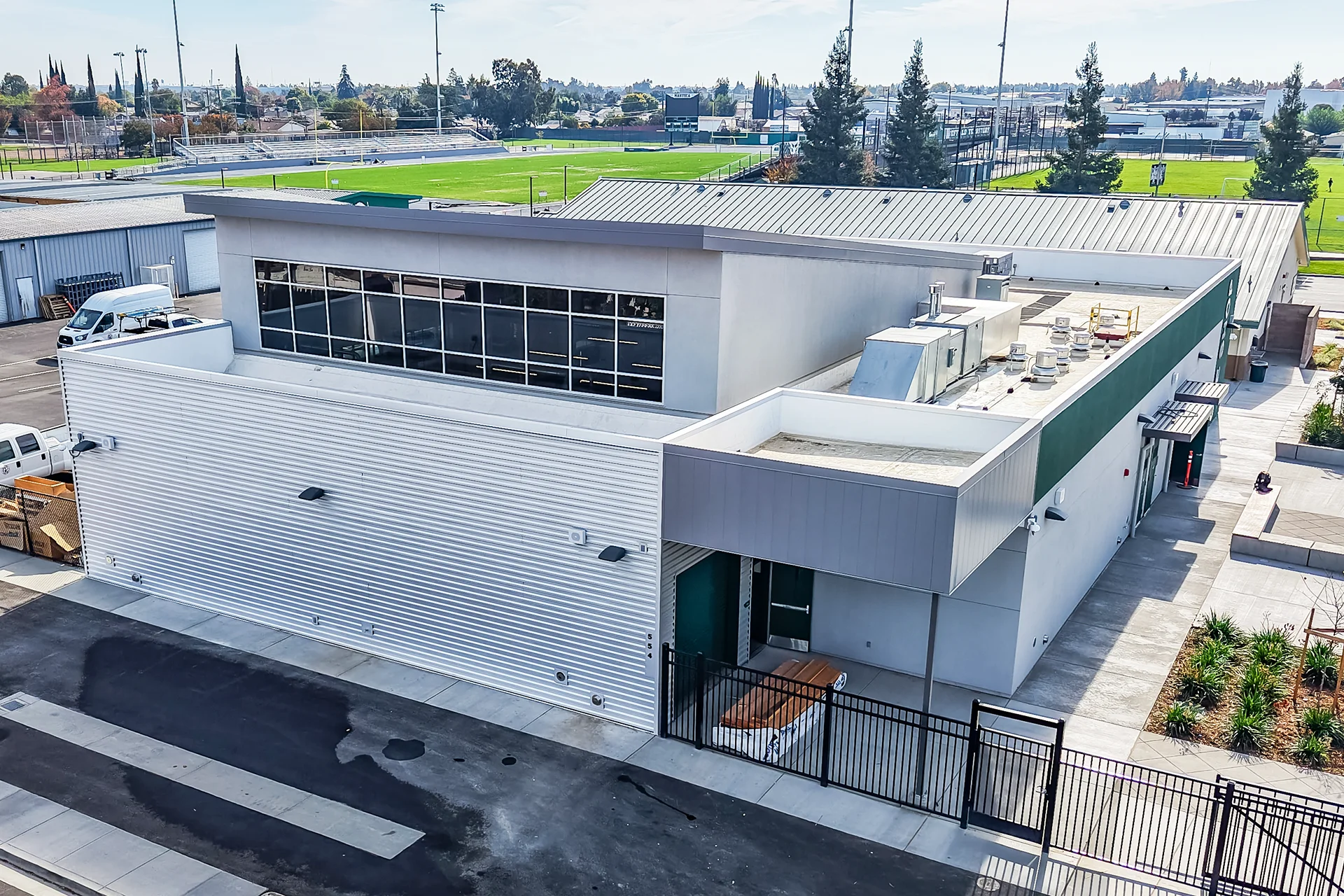 Drone view of a commercial vocational workshop structure in Manteca, California, featuring horizontal corrugated metal panel siding, a large upper clerestory window band, a flat entrance canopy over black metal security fencing, and an adjacent synthetic turf athletic field in the background.