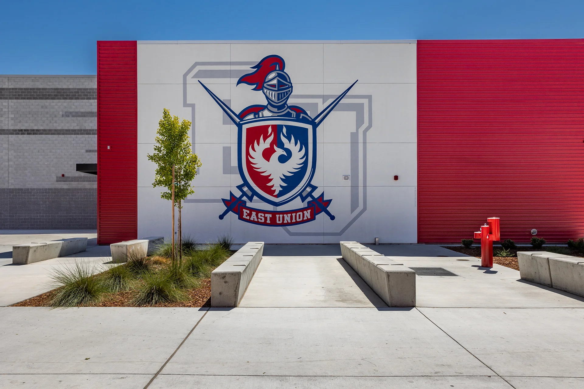 Symmetrical view of a campus pedestrian entryway highlighting geometric concrete block benches, ornamental landscaping grasses, a red utility station, and a vibrant school crest mural on a flat panel building exterior.
