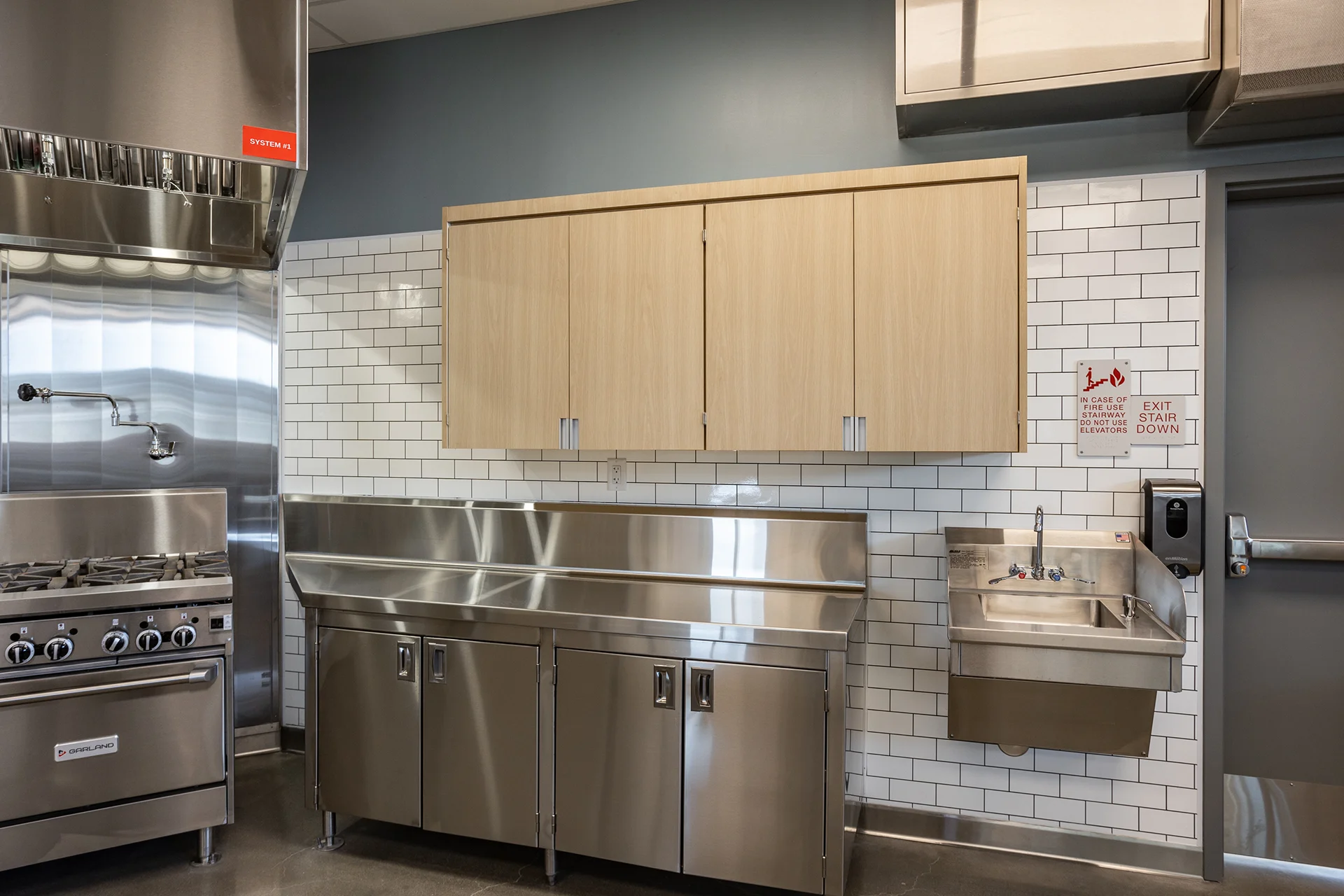 Close-up of an ADA-compliant commercial handwashing sink with splash guards and a sensor soap dispenser, adjacent to custom stainless steel cabinetry and a white ceramic subway tile wall partition.