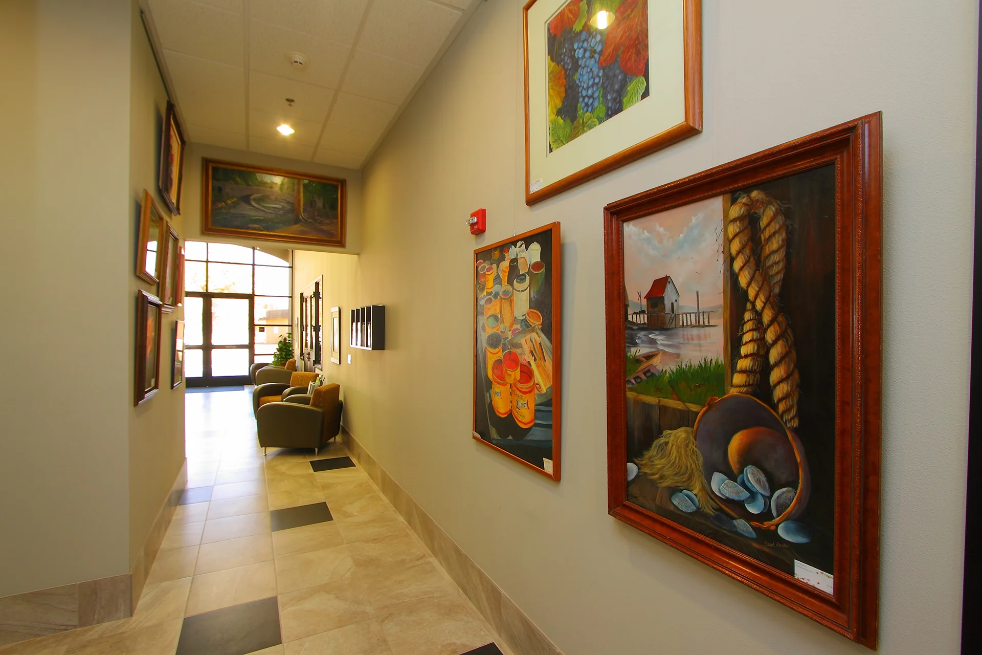 Public hallway inside Waterford City Hall featuring light neutral walls adorned with framed fine art paintings, ceramic tile flooring with dark accent inserts, leather armchairs, and an expansive glass storefront window system illuminating the far end of the corridor.