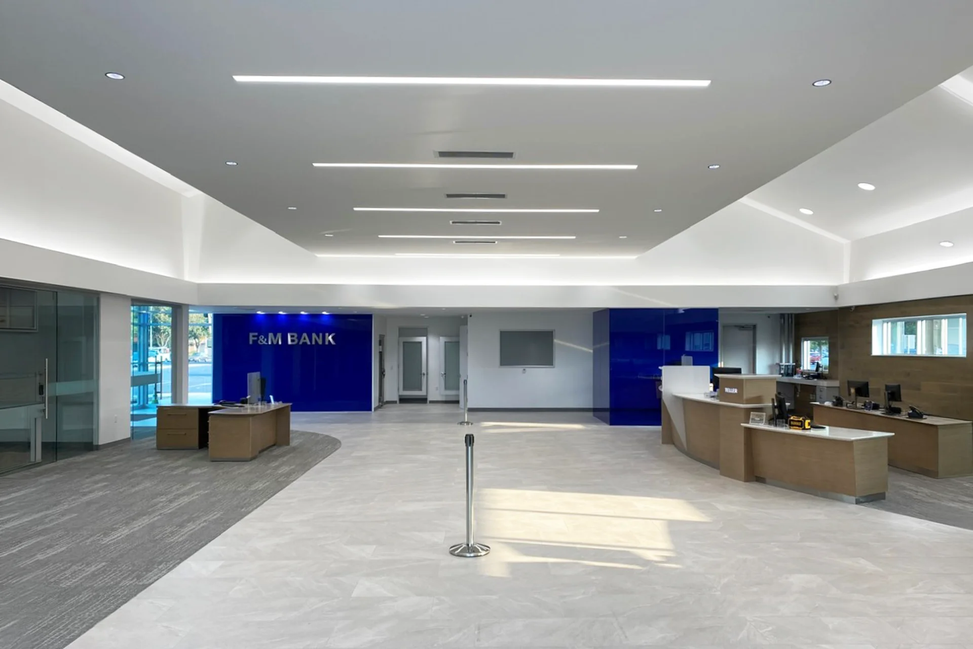 Comprehensive interior shot of the newly built F&M Bank lobby, showcasing the symmetrical layout of the main floor, mixed carpeting and tile surfaces, a blue accent wall with silver corporate branding, and modern acoustic ceiling transitions.