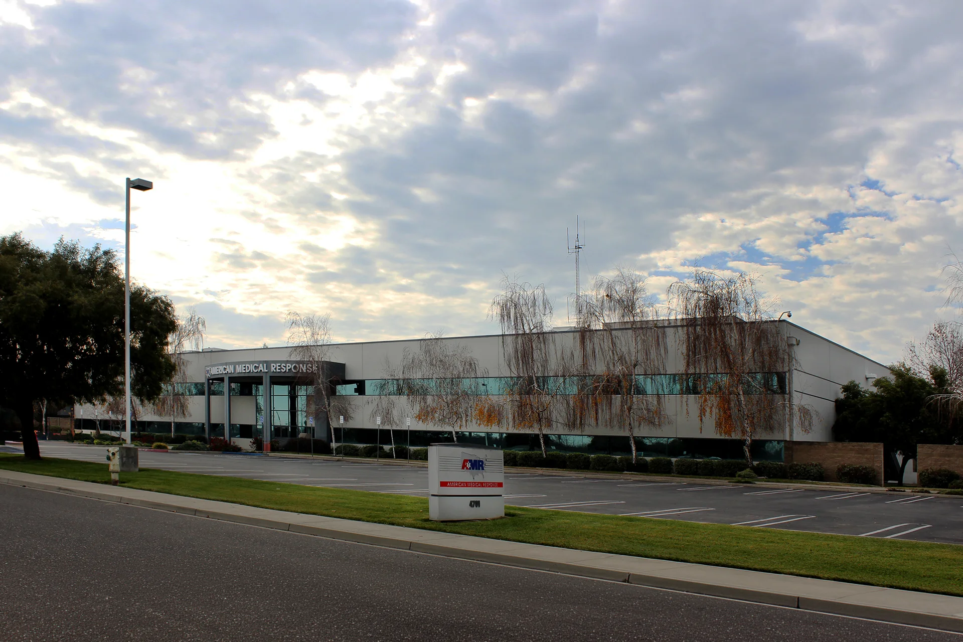 Angled landscape view of the American Medical Response facility on Stoddard Road, showcasing the commercial stucco facade, continuous tinted window bands, landscaped lawn with birch trees, and an exterior monument sign near the roadway.