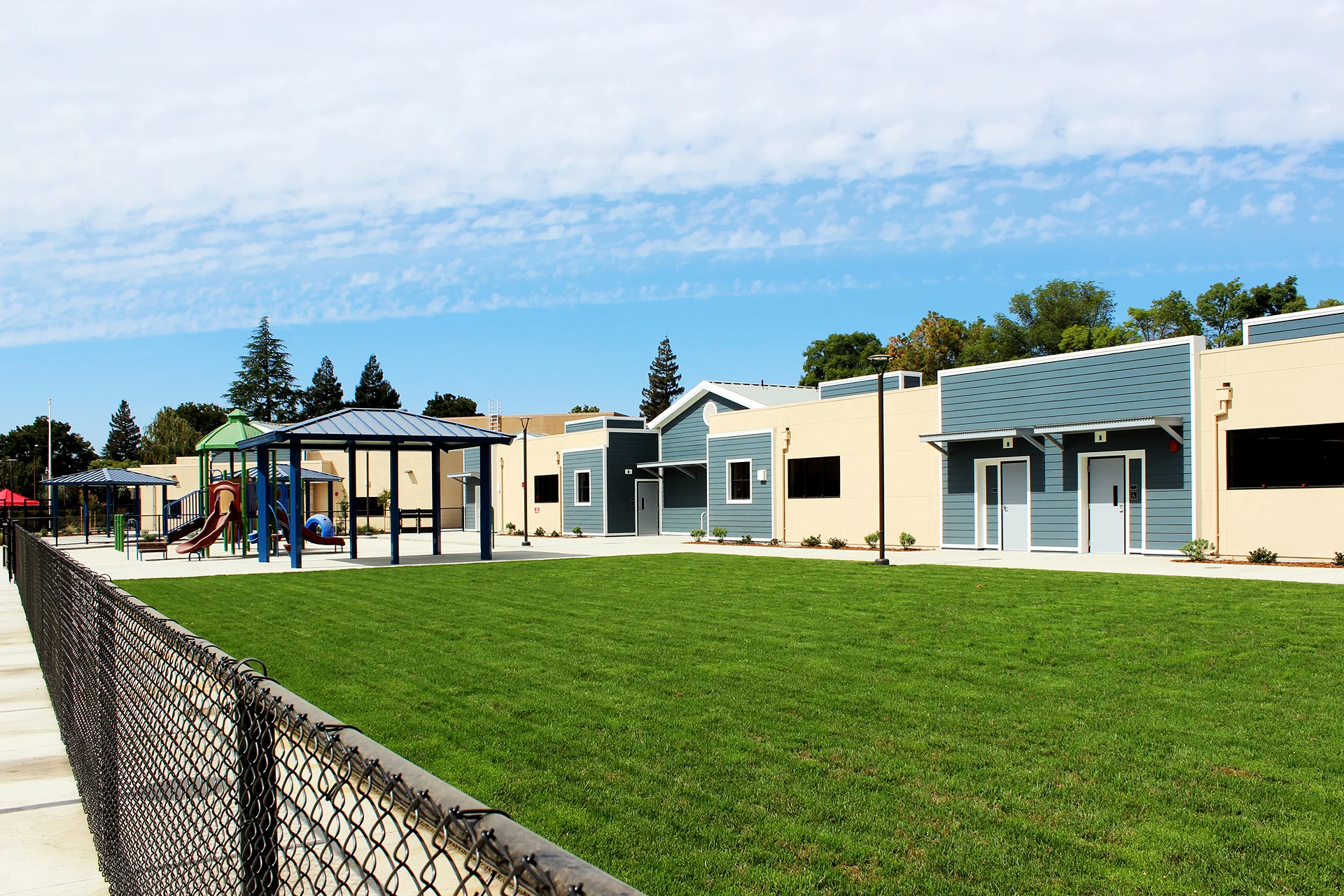 Outdoor playground and classroom wing at Quail Lakes Elementary School featuring shaded play structures, landscaped green space, covered gathering areas, and modern educational buildings.