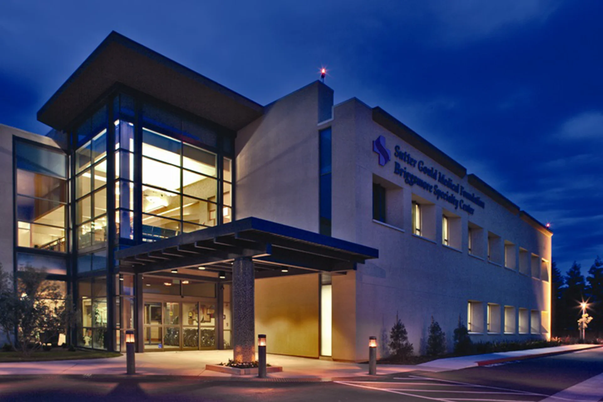 Main entrance of the Sutter Gould Briggsmore Specialty Center featuring a glass atrium, covered patient drop-off, illuminated healthcare facility, and contemporary medical office architecture at dusk.