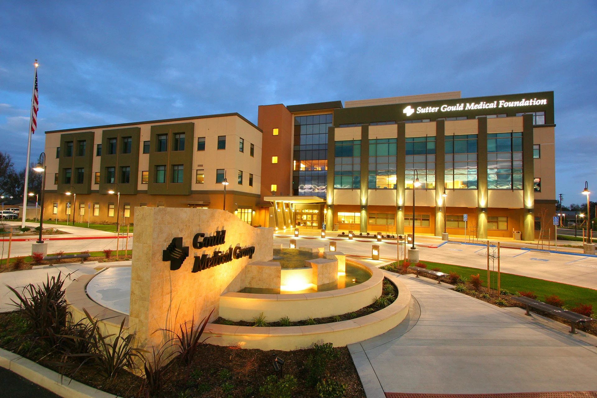 Front entrance of the Sutter Gould Medical Foundation medical office at 600 Coffee Road featuring the illuminated patient entrance, monument sign with water feature, landscaped grounds, and contemporary healthcare architecture at dusk.