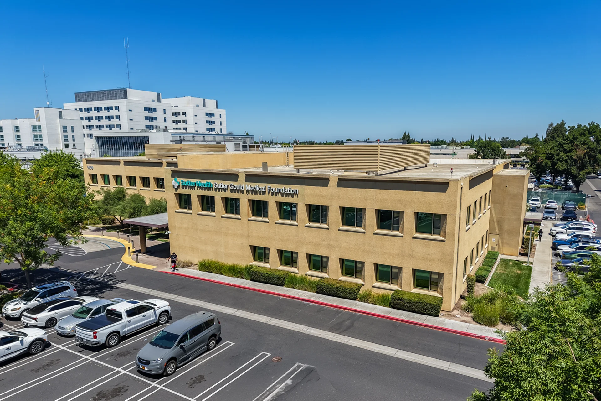 Elevated exterior view of the Sutter Gould Briggsmore Specialty Center featuring the outpatient medical building, patient parking, landscaped grounds, and the adjacent Memorial Medical Center campus in Modesto, California.
